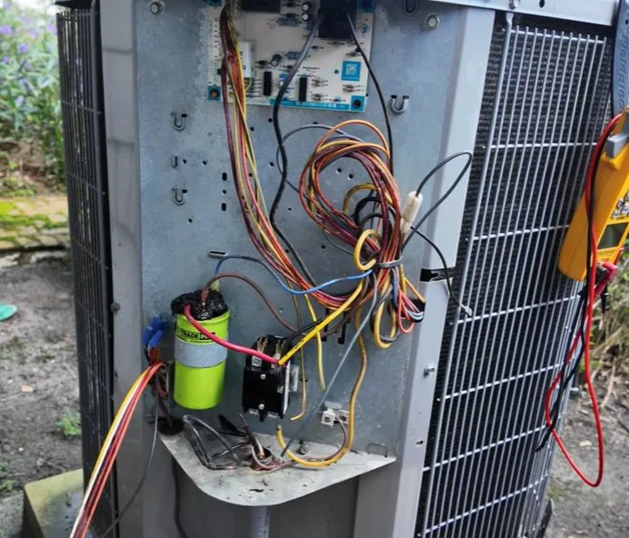Open electrical compartment of a Carrier outdoor AC unit showing the control board, wiring, a run capacitor and a contactor, with a multimeter clamped to the cabinet.