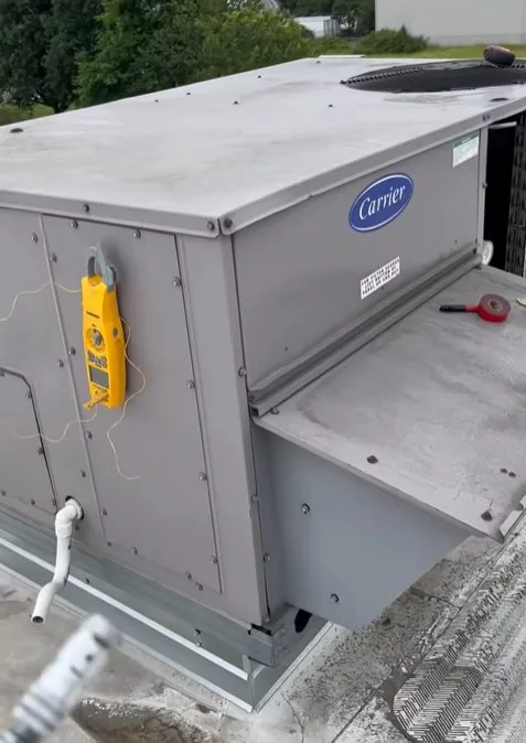Grey Carrier rooftop package unit with the Carrier badge on its side, a yellow multimeter hanging from the cabinet, and a drain line at its base.