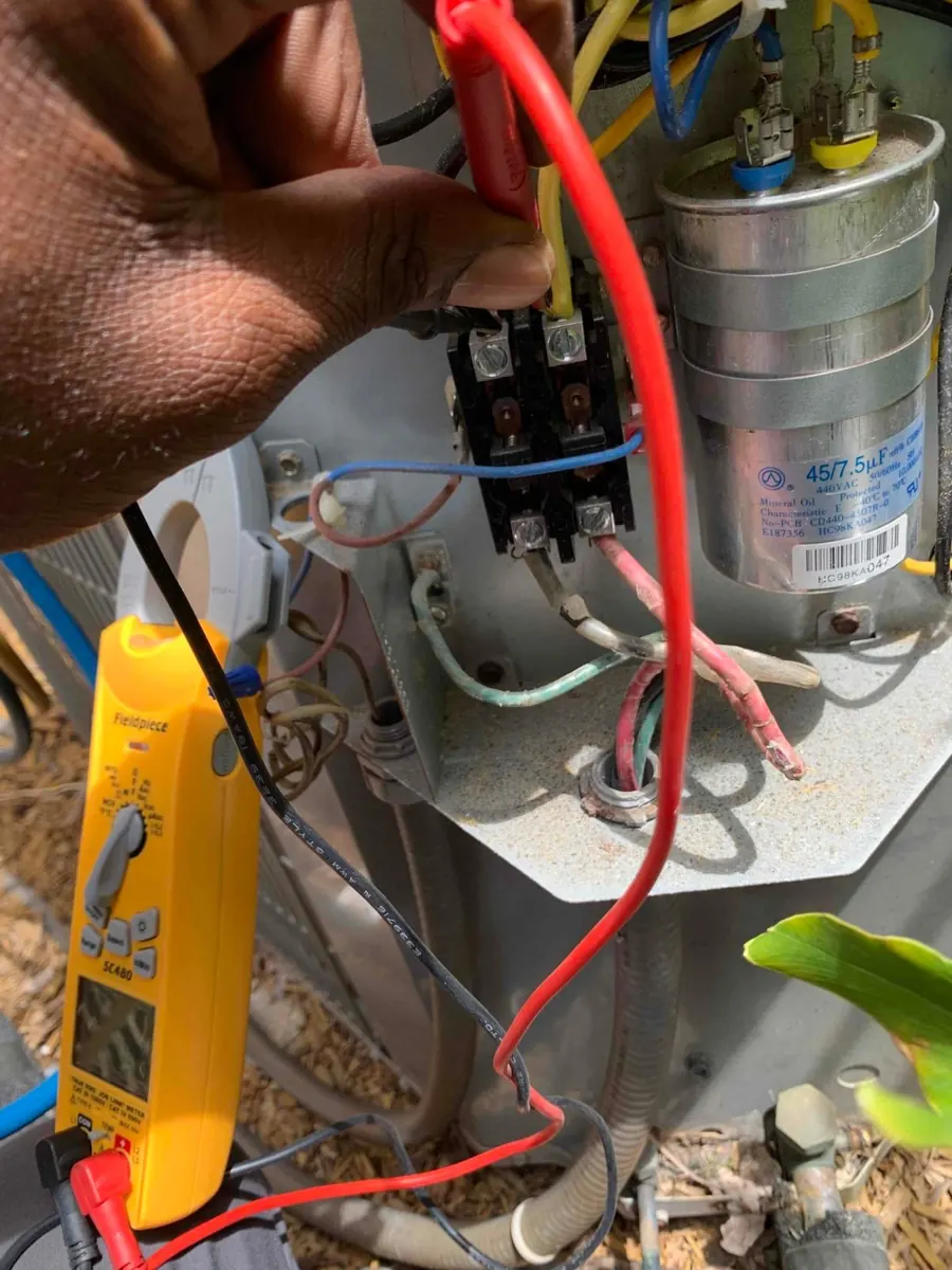 Liberty Air and Electric tech holding meter leads on a contactor inside an outdoor AC unit, with the run capacitor beside it and a clamp meter hanging below.