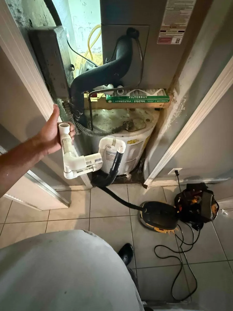 Technician holding a PVC condensate trap assembly in front of a closet air handler above a water heater, with a shop vac hose connected to the drain line.