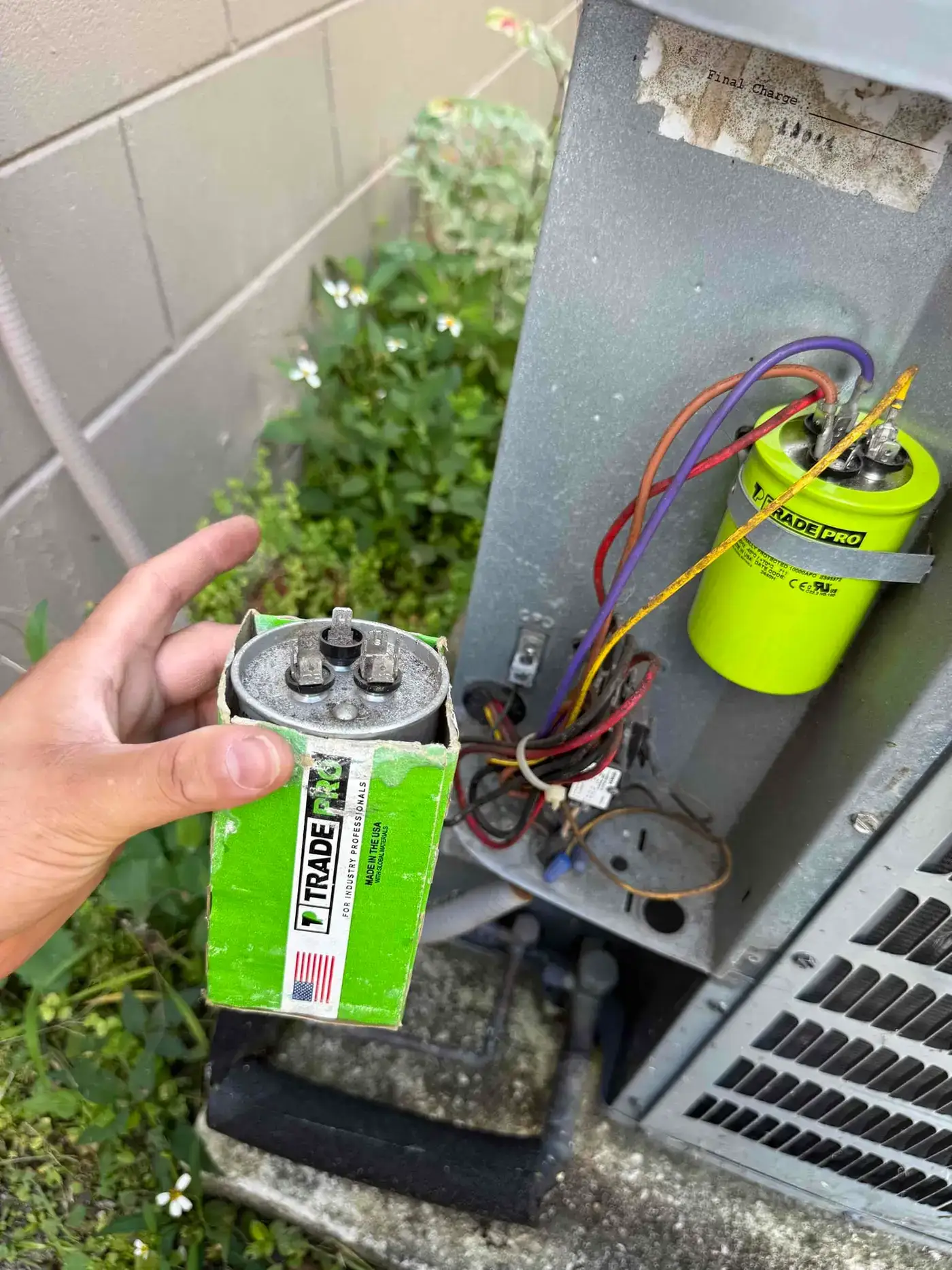 Liberty Air and Electric tech holding a new dual-run capacitor in its box next to the installed replacement inside a residential AC condenser unit in Pompano Beach, Florida.