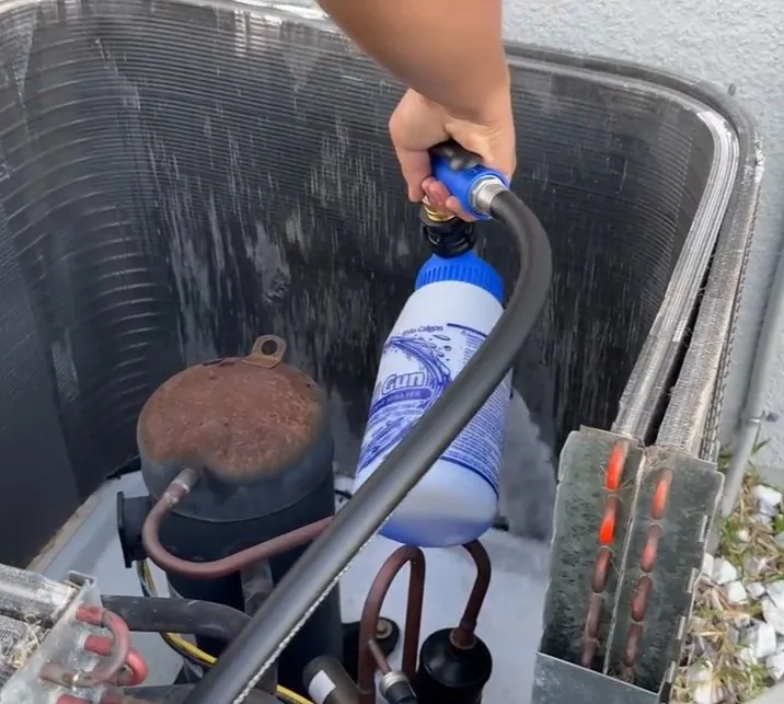 Liberty Air and Electric technician holding a hose-end sprayer inside an outdoor AC unit, above the compressor, with the condenser coil along the cabinet walls.