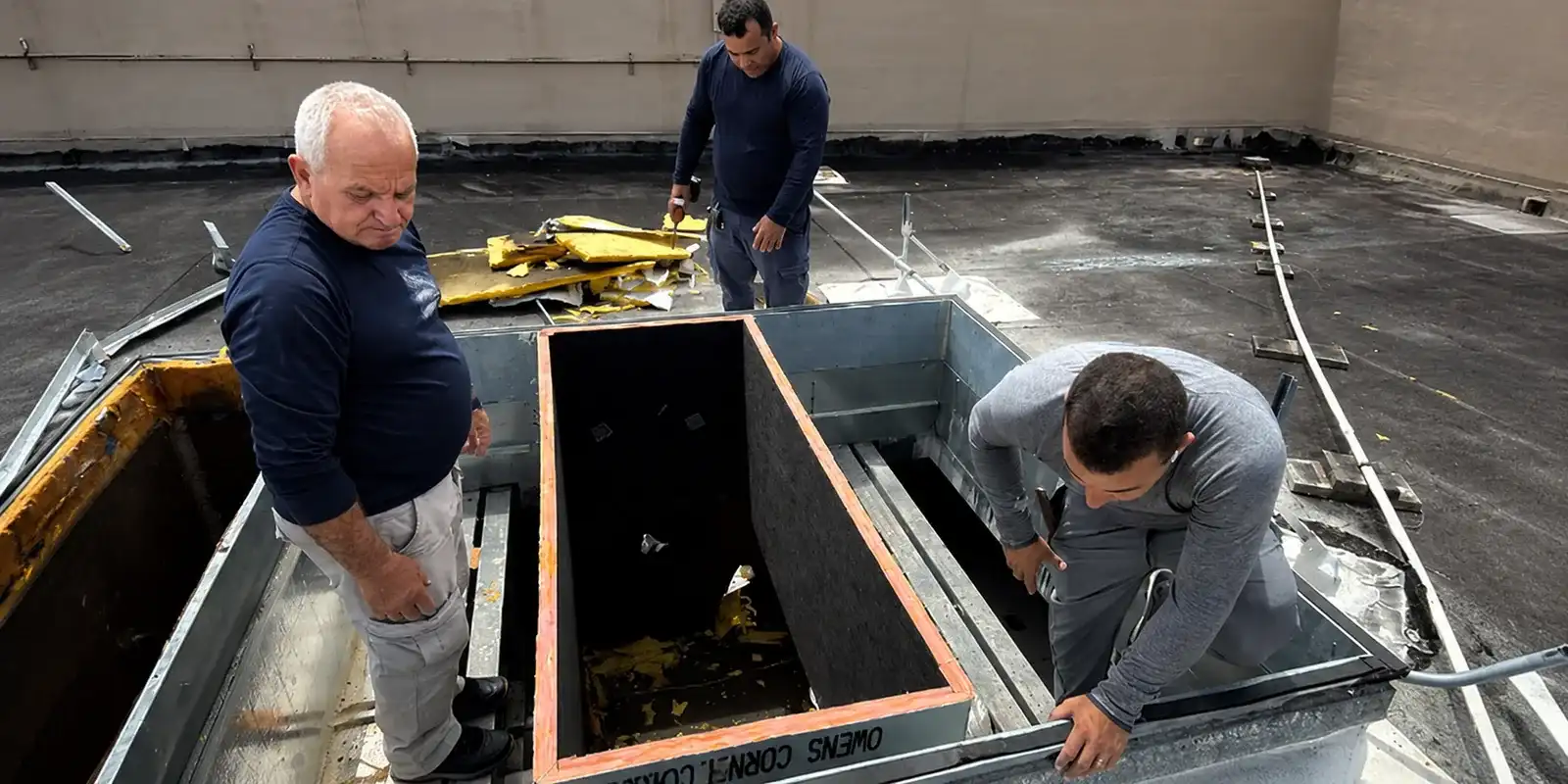 Three Liberty Air and Electric techs working a commercial rooftop during a re-roof project, gathered around a curb opening with the roof opened down to the insulation layer.