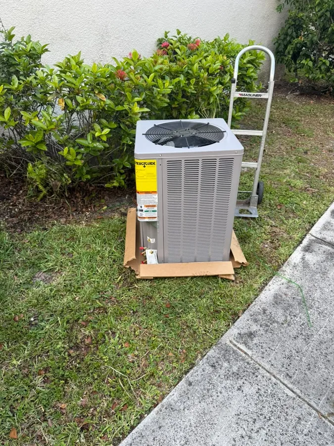 Grey outdoor AC condenser with a top fan grille and louvered side panels, sitting on flattened cardboard on the grass beside a sidewalk, with a yellow EnergyGuide label on its corner and a hand truck parked by a flowering hedge.