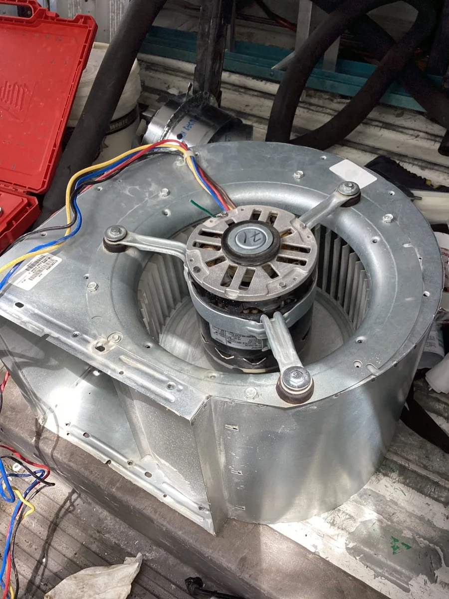 Galvanized blower housing sitting out of its cabinet on a work surface, with the blower motor mounted in the center of the wheel on a three-arm bracket, colored wiring leads running off the motor, and a red tool case beside it.