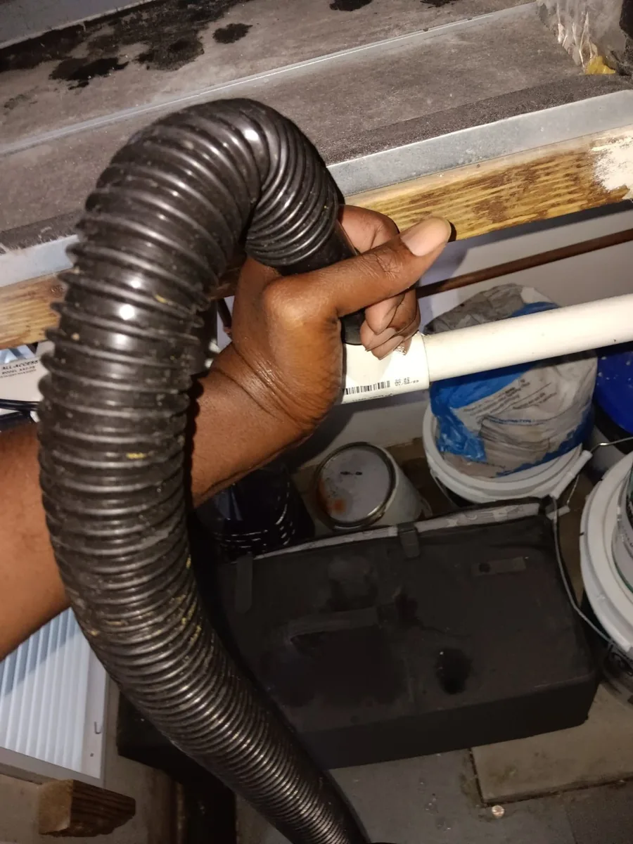 Liberty Air and Electric technician holding a wet/dry vac hose over the end of a white PVC drain line, with buckets and paint cans stored below.